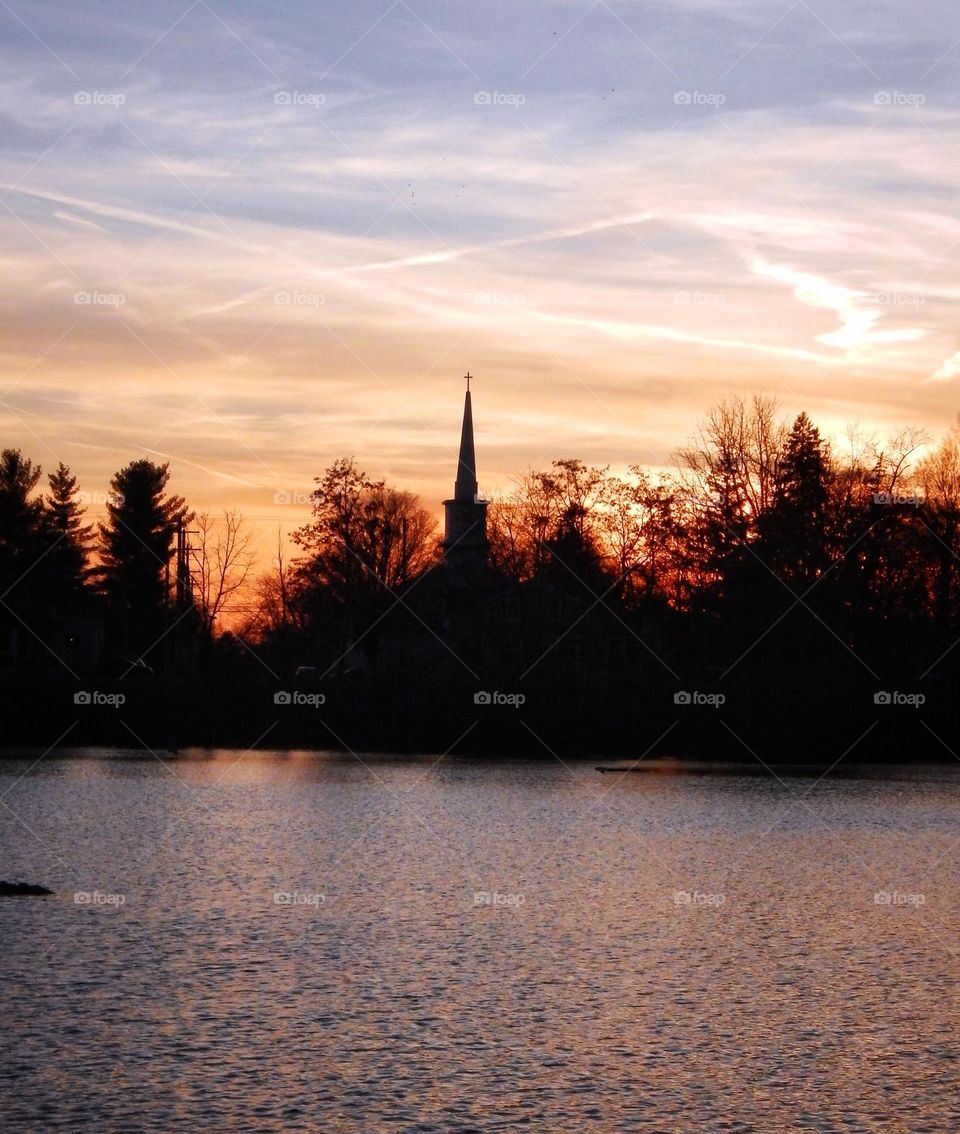 Steeple of a church on a riverbank hidden within the shadows of trees as the sun sets making the sky glow orange in Michigan on Grand River