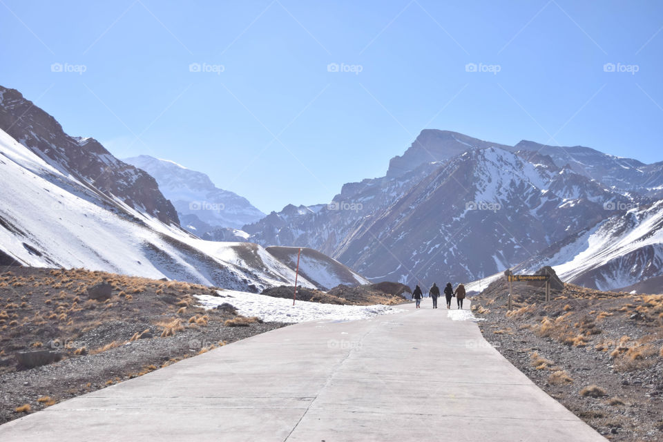 4 pessoas andando ao longe, numa trilha, por entre as montanhas da Cordilheira dos Andes. O céu está bem azul e as montanhas cheias de neve.