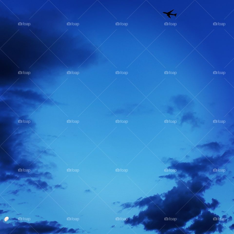 A perfect blue sky with the moon peaking behind the clouds and the silhouette of a jet flying by.