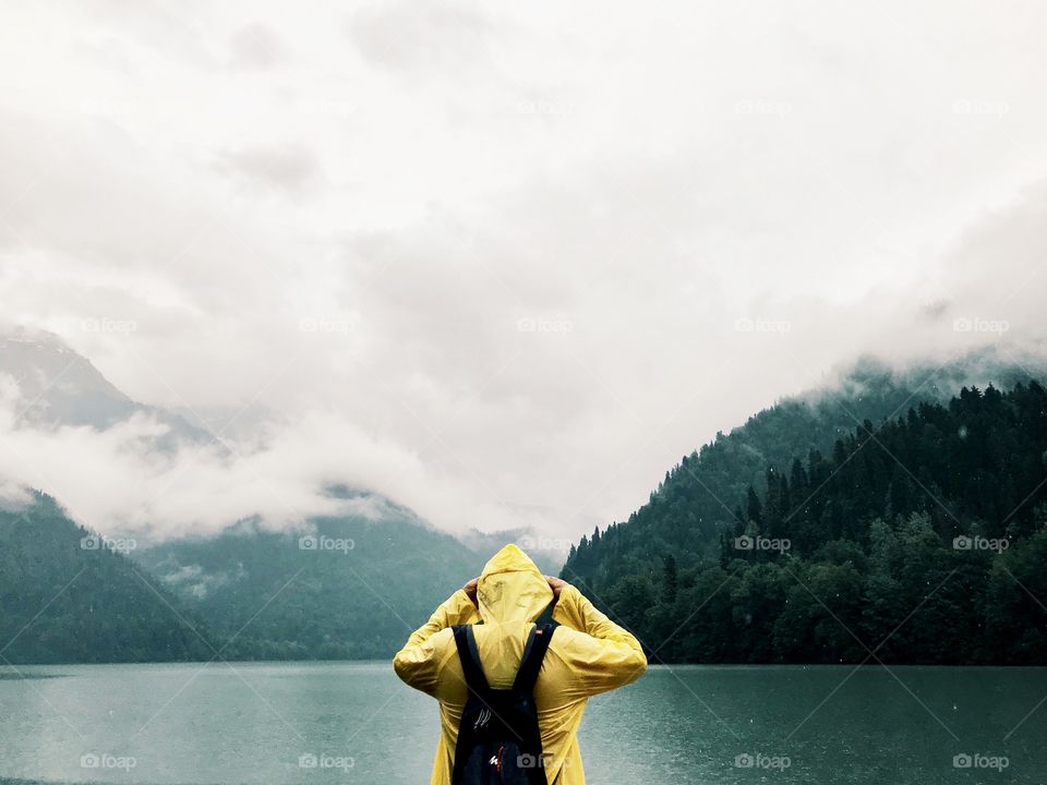 A man in a yellow cloak stands with his back against the background of beautiful mountains, lakes, clouds. Travel, nature