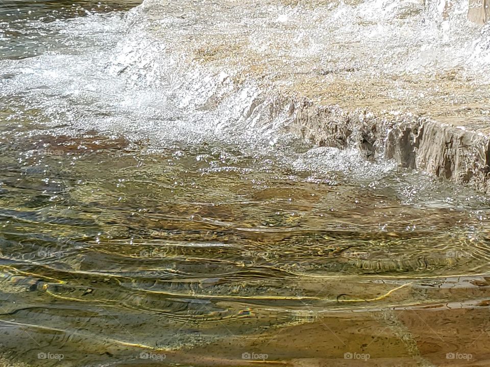 Closeup of a water fountain that fills an urban Spring fed pool with water at a city park. The surface appears to be made out of flagstone and local limestone.