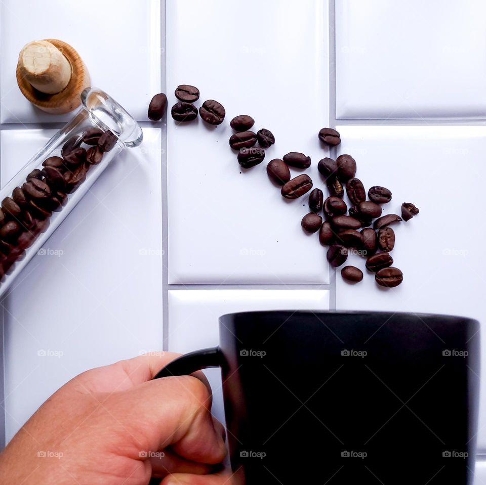 Coffee beans spilling out of slender glass container into a black coffee mug being held by a hand. This is on a vertical white kitchen tile background.