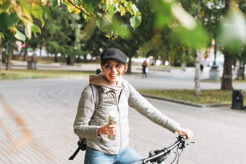 Portrait of fashionable young pretty woman in cap and sunglasses on bicycle with thermo mug on sunny autumn day in city park
