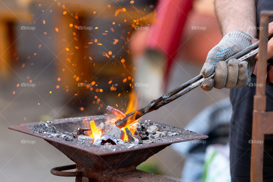 Closeup of blacksmith hand holding tongs with metal piece and heating it in fire before forging, selective focus. Blacksmithing concept.