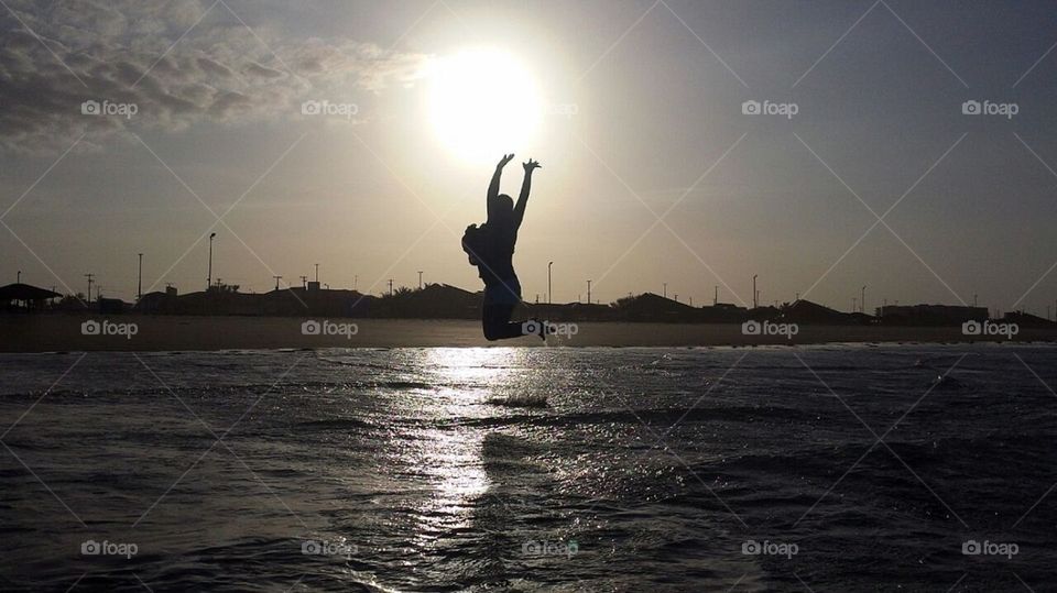 silhueta de uma mulher pulando sobre as águas do mar. Ao fundo o sol se pondo.