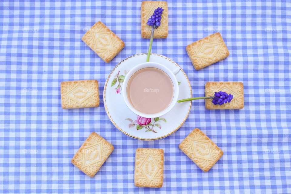 A cup of tea with biscuits in the shape of a clock on a gingham tablecloth 