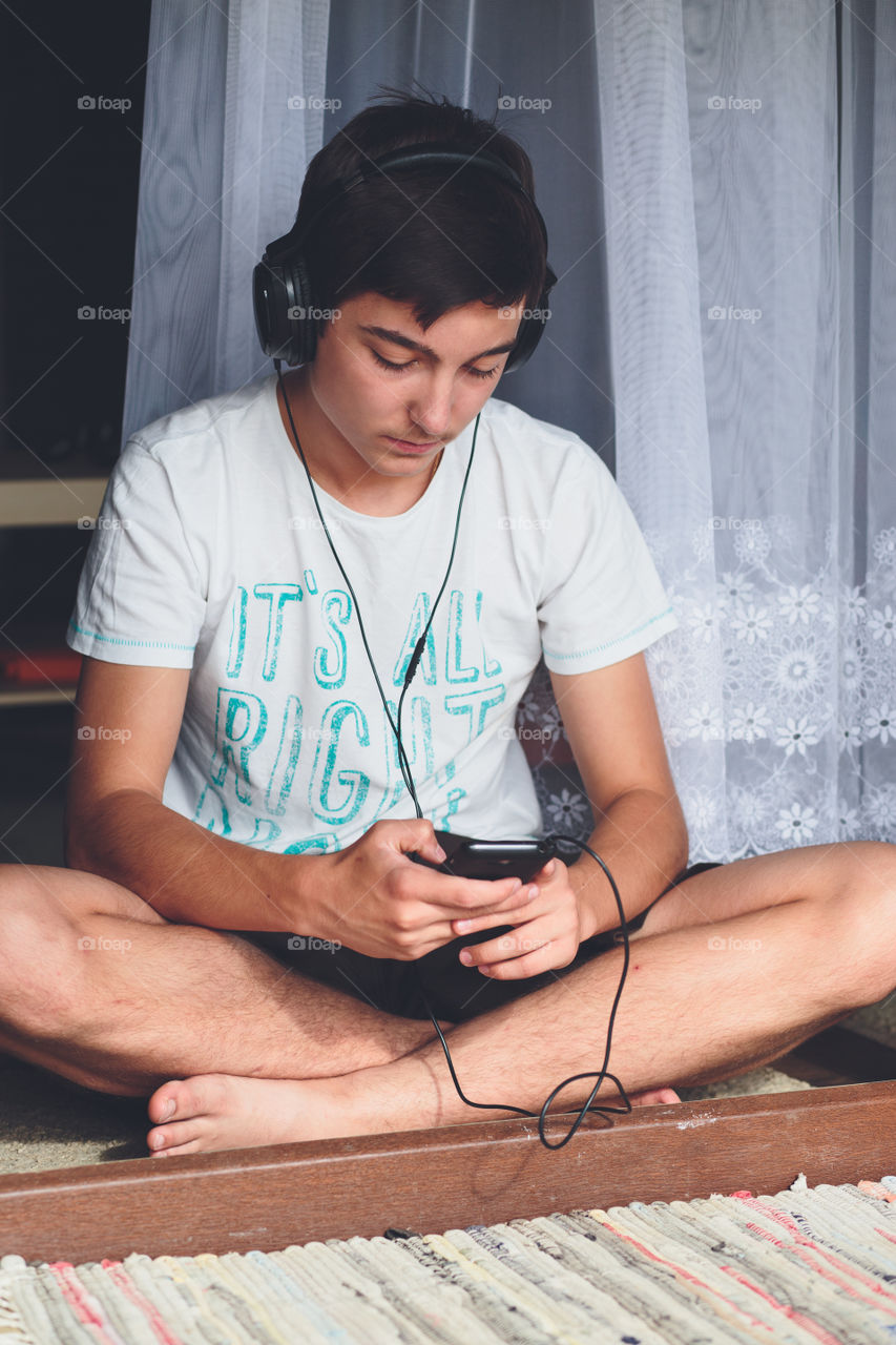 Concentrated on mobile phone. Young boy sitting at the patio door and listening to music from mobile phone