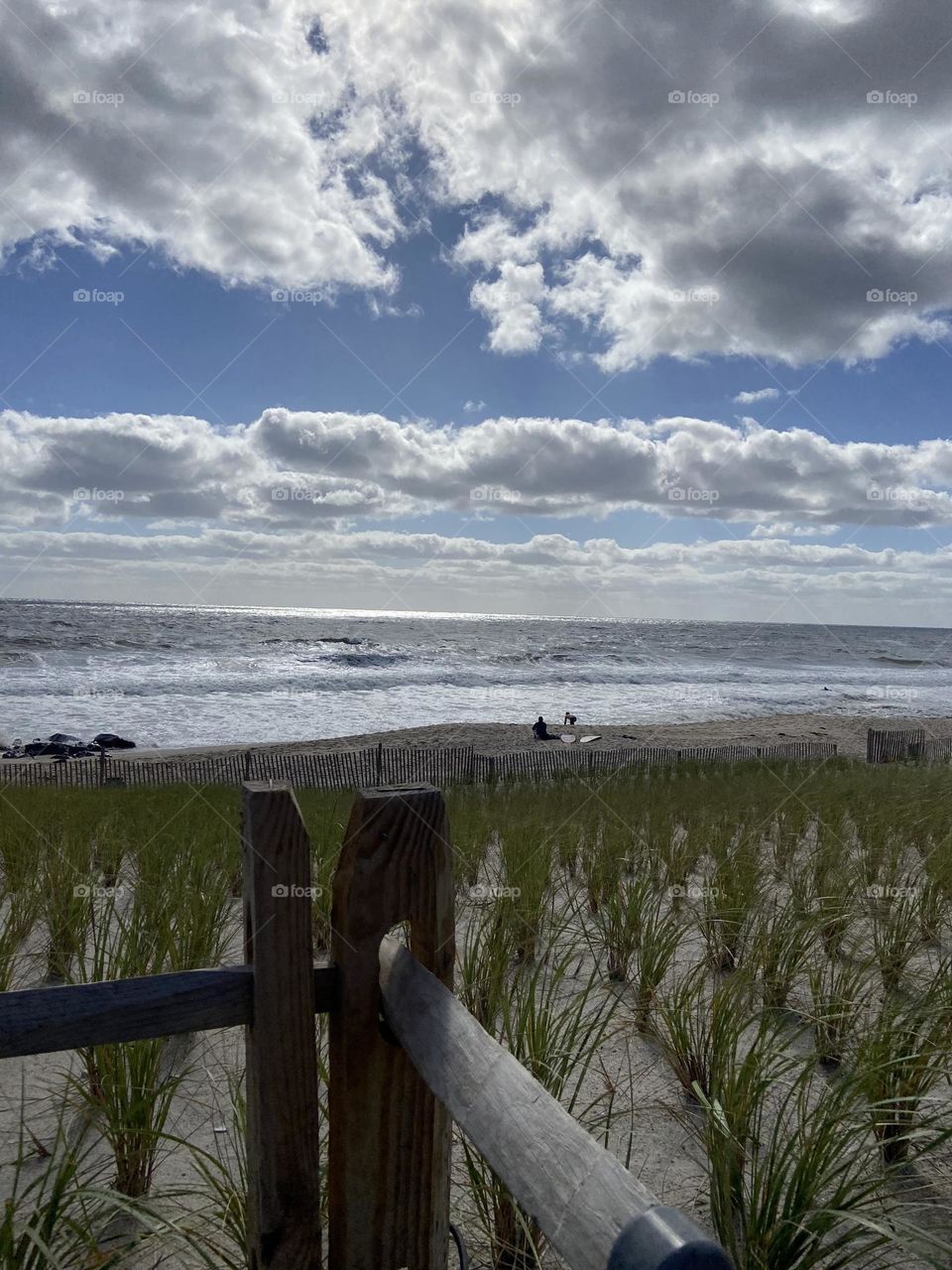 The grass-covered dunes on the beach in Bay Head, NJ. Taken from a platform you reach by stairs that offers an overhead view. Clouds fill the sky above, but in between are patches of bright blue. A few people are on the beach enjoying the ocean. 