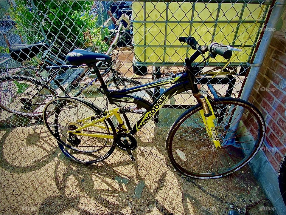 Seen leaning up against a yellow plastic pane and a metal grid are several bicycles as well as bicycle parts on a bright sunshiny day in the summer of 2022. Hypnotic Productions