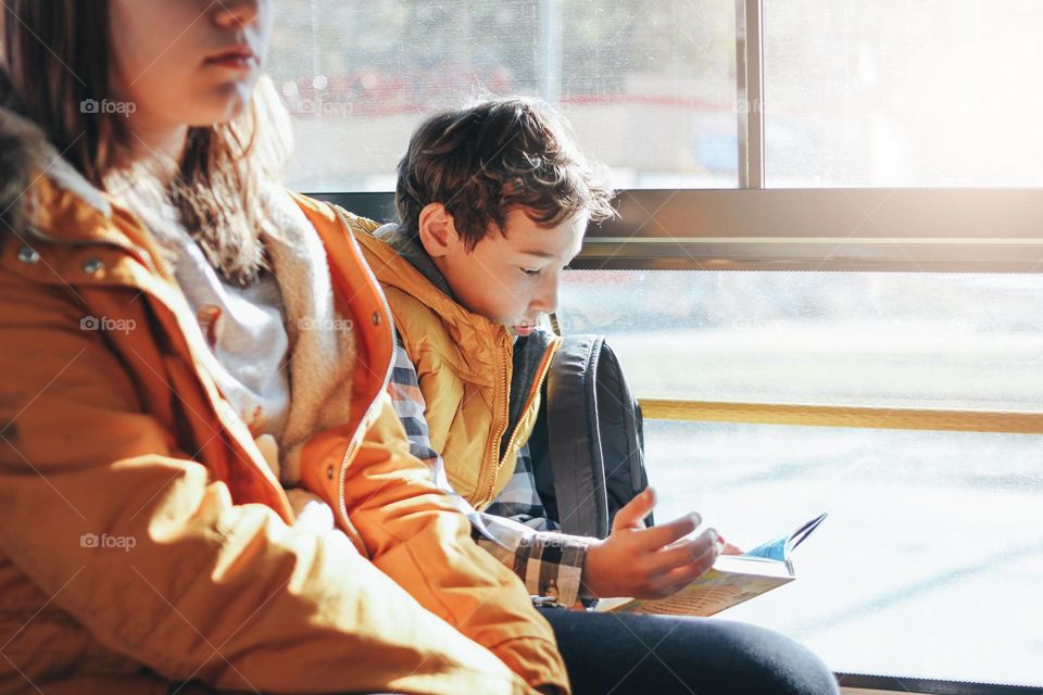 Children students in yellow jackets riding in the city bus. Boy reading a book at the window