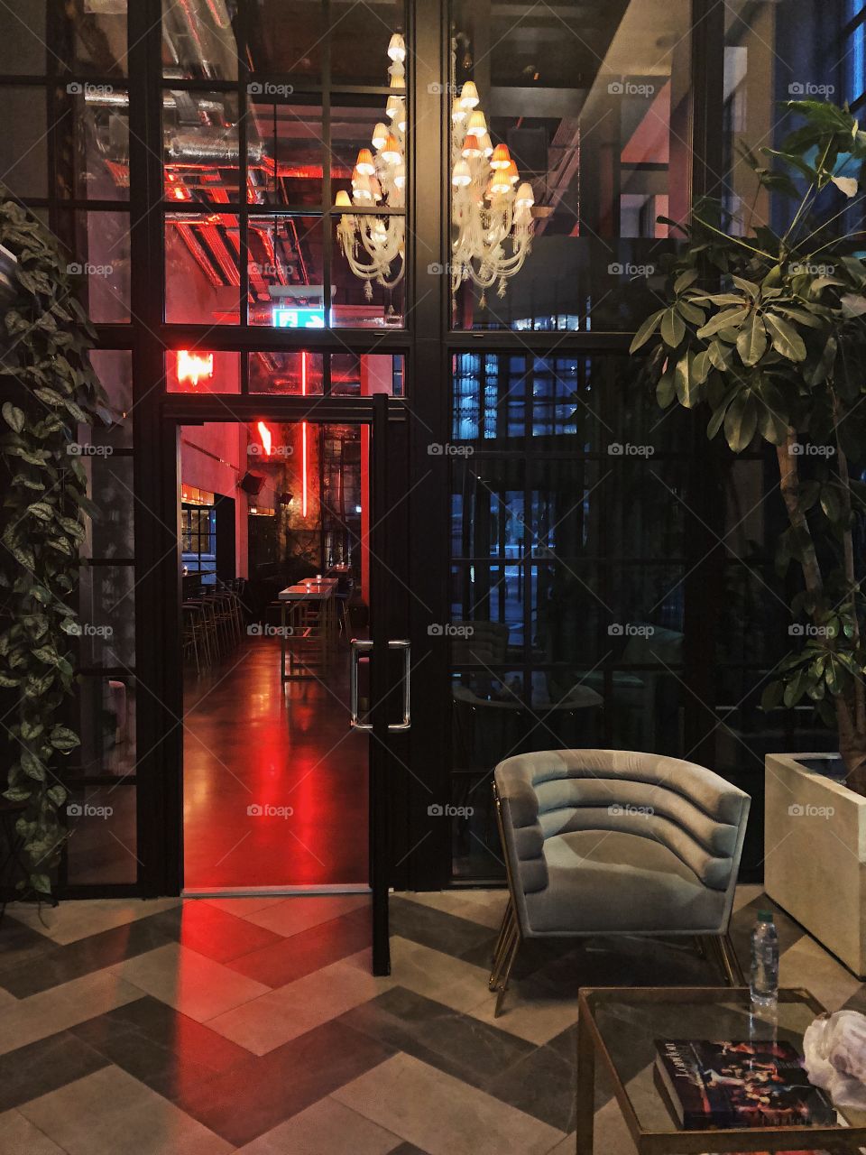 Entrance to trendy bar in east London with grey velvet armchair, modern chandelier, monochrome floor tiles and red neon lights in the background. Indoor plants feature