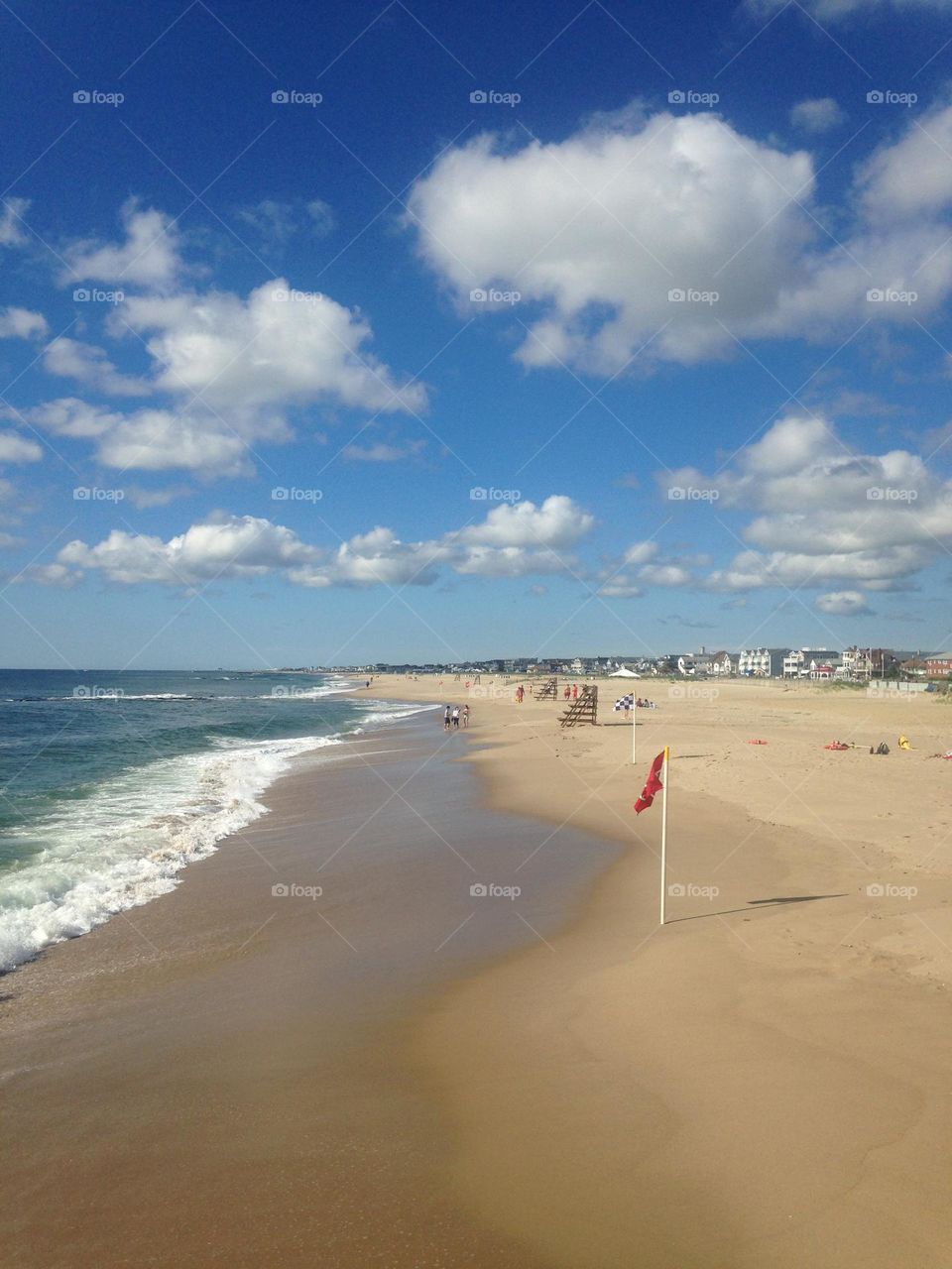A view of the ocean and beach from a pier in Ocean Grove, NJ on a Spring day. This shot is looking toward the neighboring town of Bradley Beach. Puffy white clouds accent a bright blue sky, and waves are just cresting the sand. 