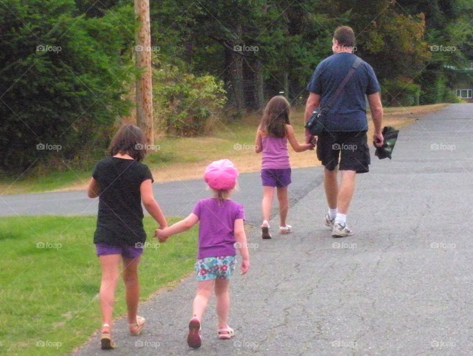 A shot of the backs of Dad and his 3 daughters taking a walk downtown to walk on the pier and maybe go for ice cream. Mom is following behind to take the picture. They’re all holding hands and enjoying the warm sunny summer day. Family time!