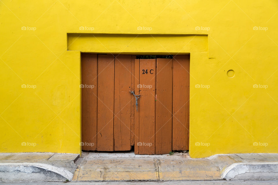 Brown wooden door on a yellow wall. Closed with chain and lock. Bright yellow wall. Sidewalk