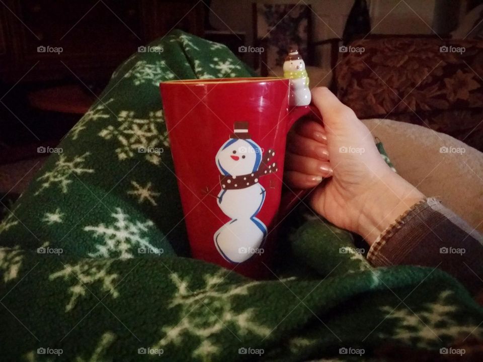 Winter Beverage in a tall mug with a snowman being held by a womans hand wrapped in a cozy green blanket at home