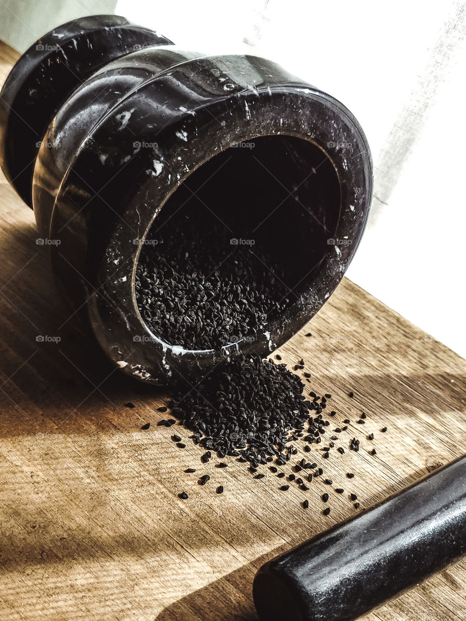 A black stone mortar tipped over with medicinal black seeds nigella sativa falling out onto the wood surface that is also illuminated by sunlight peering through the window and casting shadows on to the wood surface.