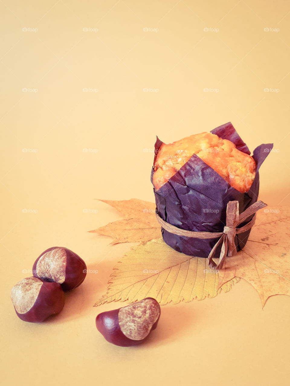 Autumn still life with pastries.  Muffin in brown baking paper with chestnuts on autumn leaves on a plain beige background.  Beginning of autumn