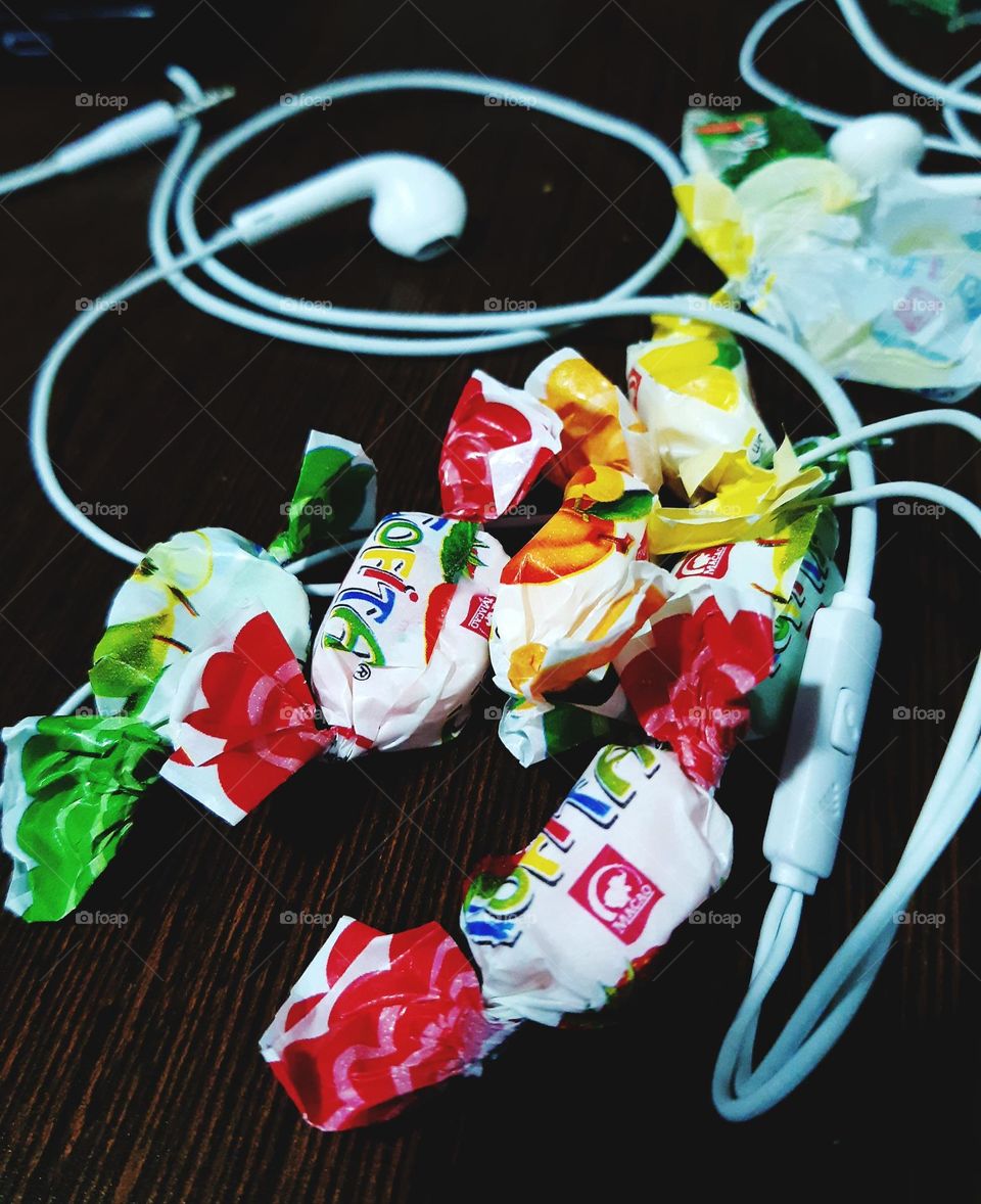 A close-up photograph captured on March 22, 2024, showcasing colorful candy wrappers scattered across a wooden surface, accompanied by white earphones. The vibrant composition blends everyday objects with playful colors, creating a casual
