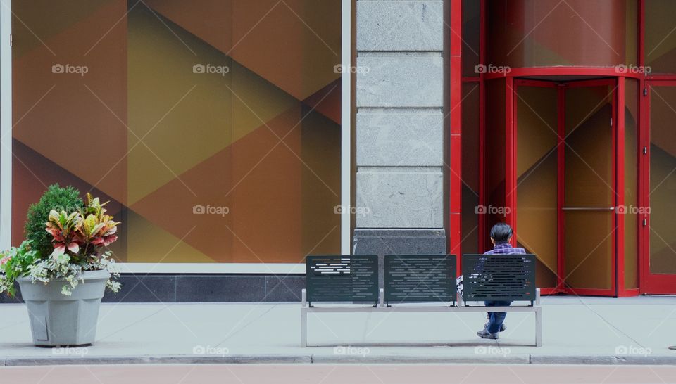 A back view of a man seated on a street bench facing a building in Manhattan, NYC.