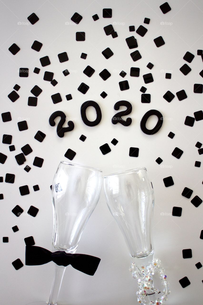 Conceptual overhead view of champagne glasses representing a man and a woman with the year ‘2020’ in black with black confetti on a white background 