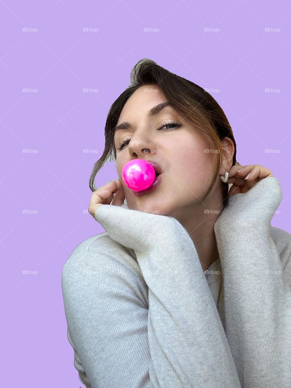 The image shows a woman wearing a light gray sweater, blowing a bright pink bubble gum bubble. She is posing with her hands resting near her chin, and the background is a pastel lavender color.