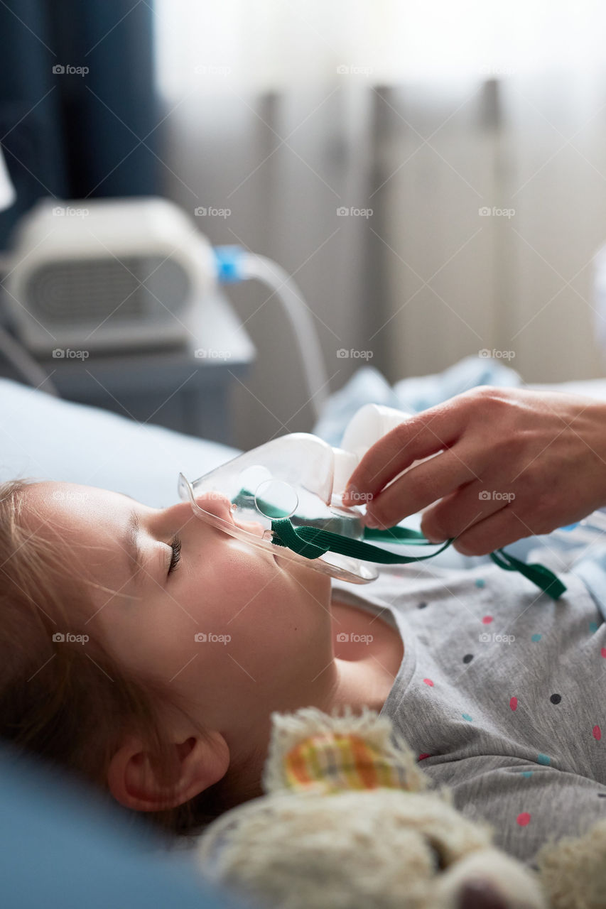 Sick little girl having medical inhalation treatment with nebuliser. Child with breathing mask on her face lying in bed