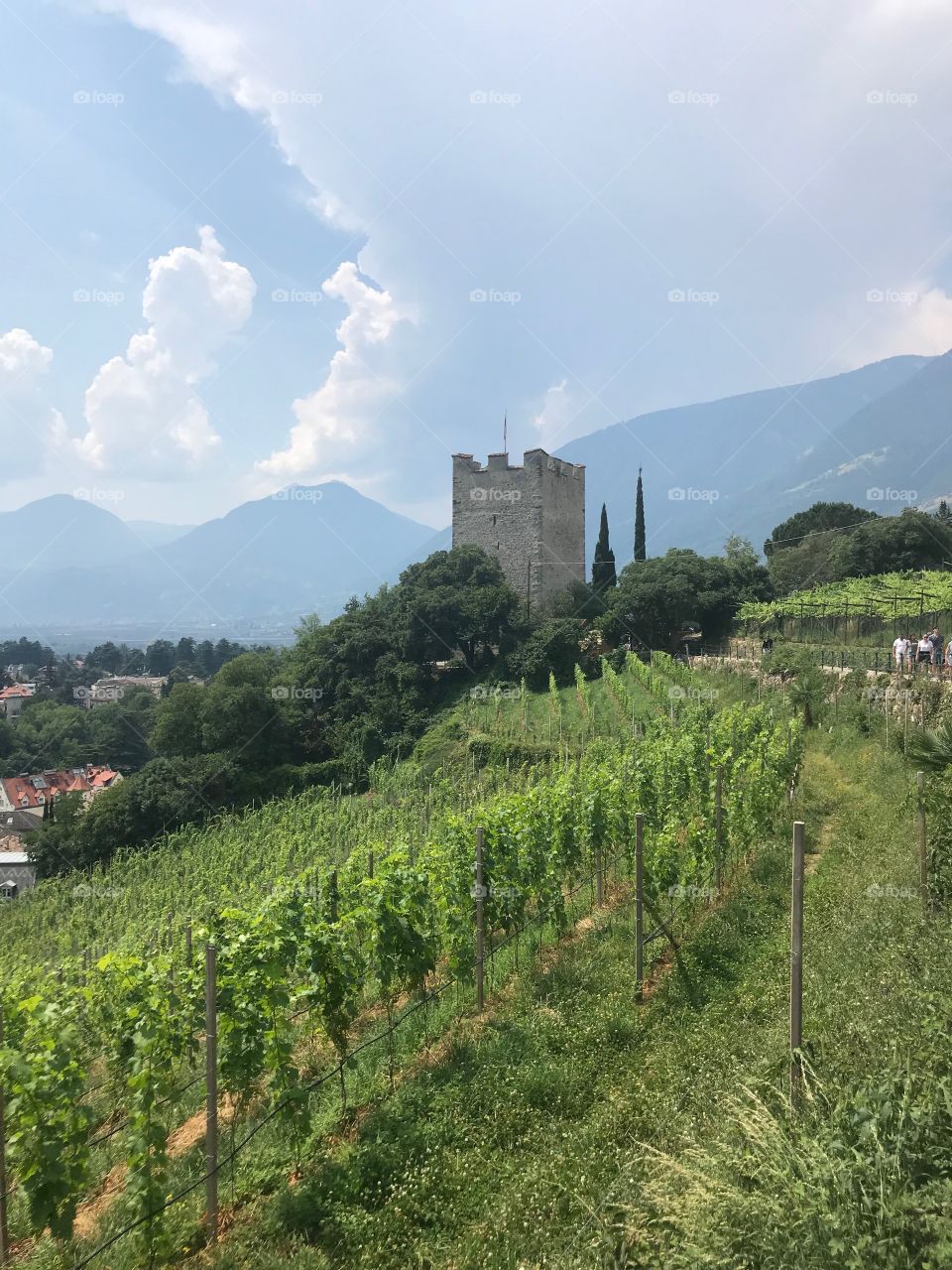 a citadel in the middle of the lush natural landscape , surrounded by vineyards and trees, there are some people walking on the small road above , red roofs in the canopy, few mountains, big clouds on the sky 