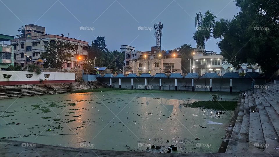 This is a picture of a beautiful pond in the evening in Basunagar Madhyamgram. The pond edge is surrounded by concrete stairs. The pond is filled with water plants. Light reflection is shown in the pond. Some high-rise buildings and trees are shown.