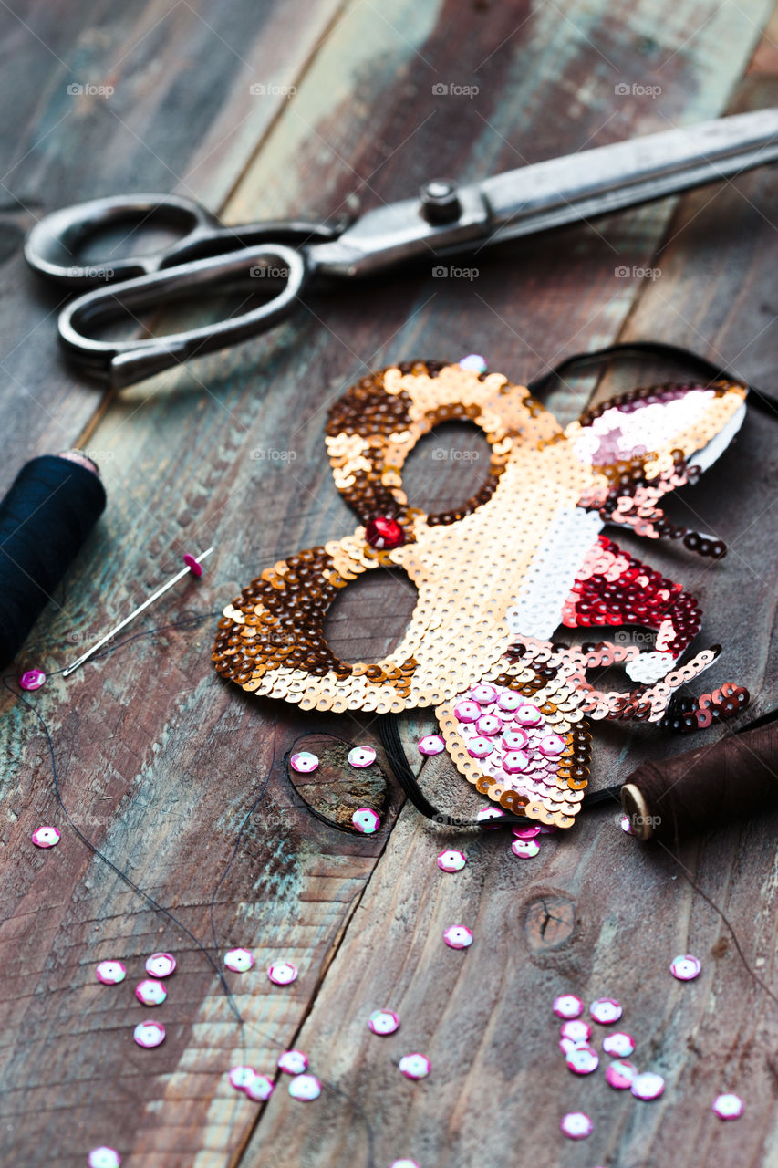 Close shot of female hand decorating Christmas reindeer mask with sequins using thread needle and  scissors following diy ideas on wooden table