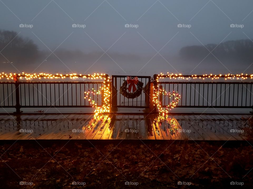 Nighttime holiday lights adorn a fence and gate that overlooks a foggy lake during dusk