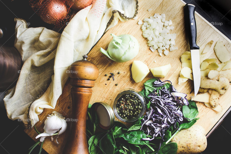 Elevated view of cozy home kitchen cooking and preparing fresh market vegetables on wood cutting board with green peppercorns, spinach, onions, chopped cabbage, kohlrabi, garlic with pepper mill, knife and table linens healthy food art photography