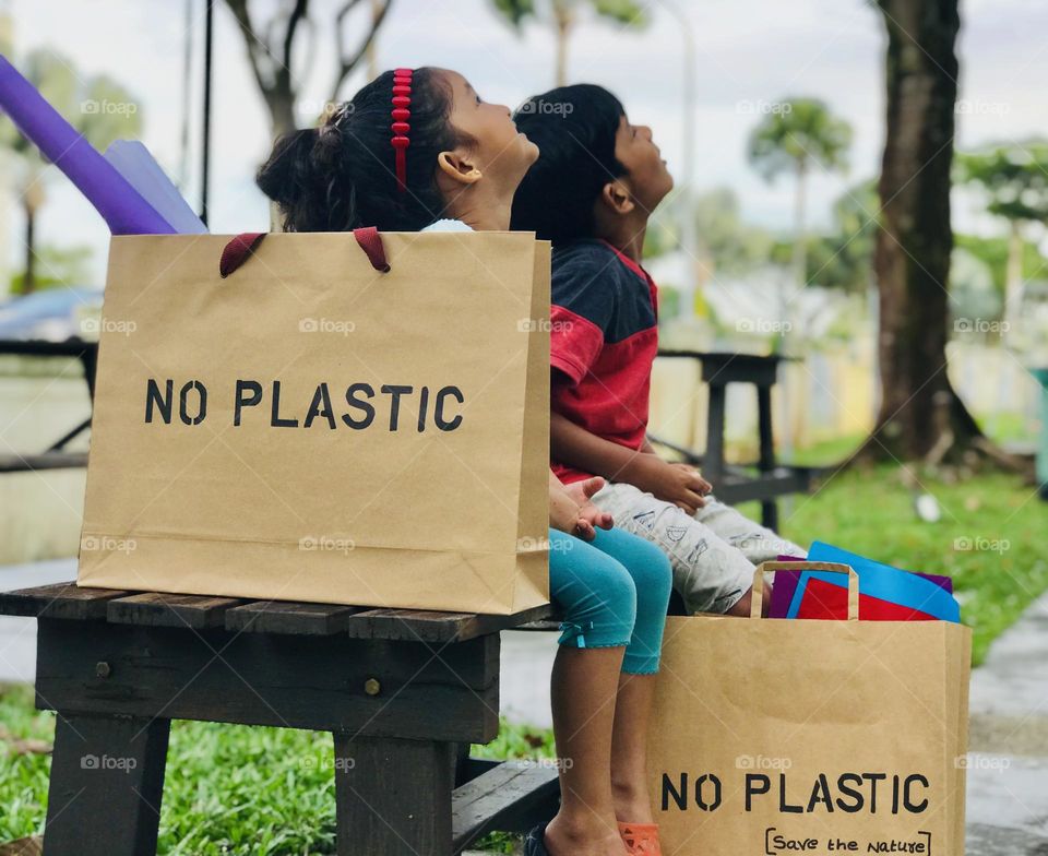 Use paper bags to reduce plastic production. Here one boy and girl put the paper bagels beside them which wrote no plastic ( save nature )on it and looking upwards. Kids encouraged to carry the paper bags instead of plastic bags.