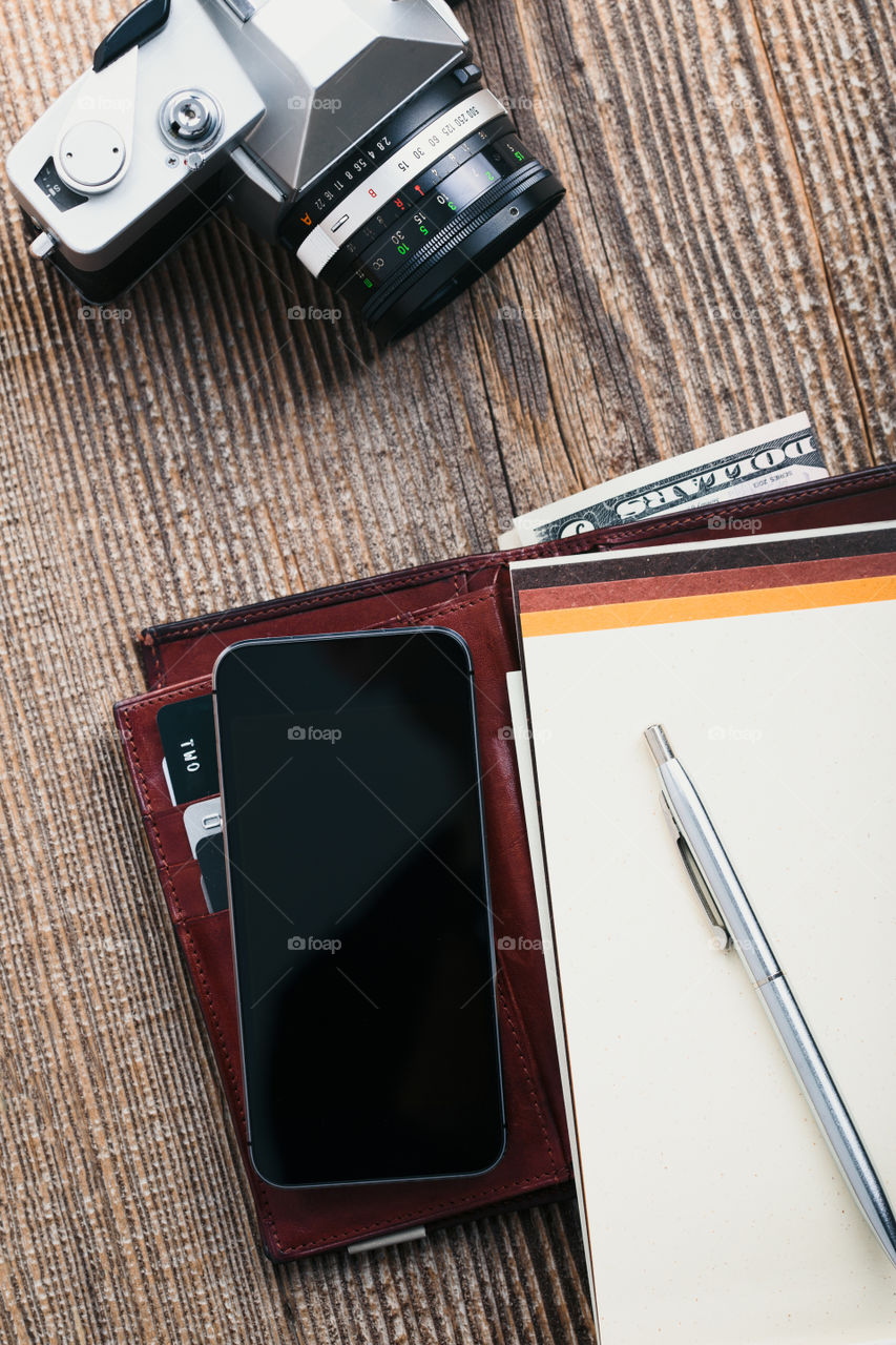 Smartphone with blank screen, camera, wallet, dollar banknotes, debit credit cards and notebook on wooden table. View from above. Portrait orientation