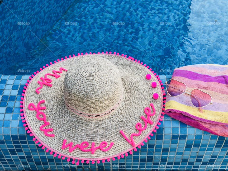 Summer hat with the words “wish you were here”,sunglasses and a pink towel on the edge of a pool with turquoise water