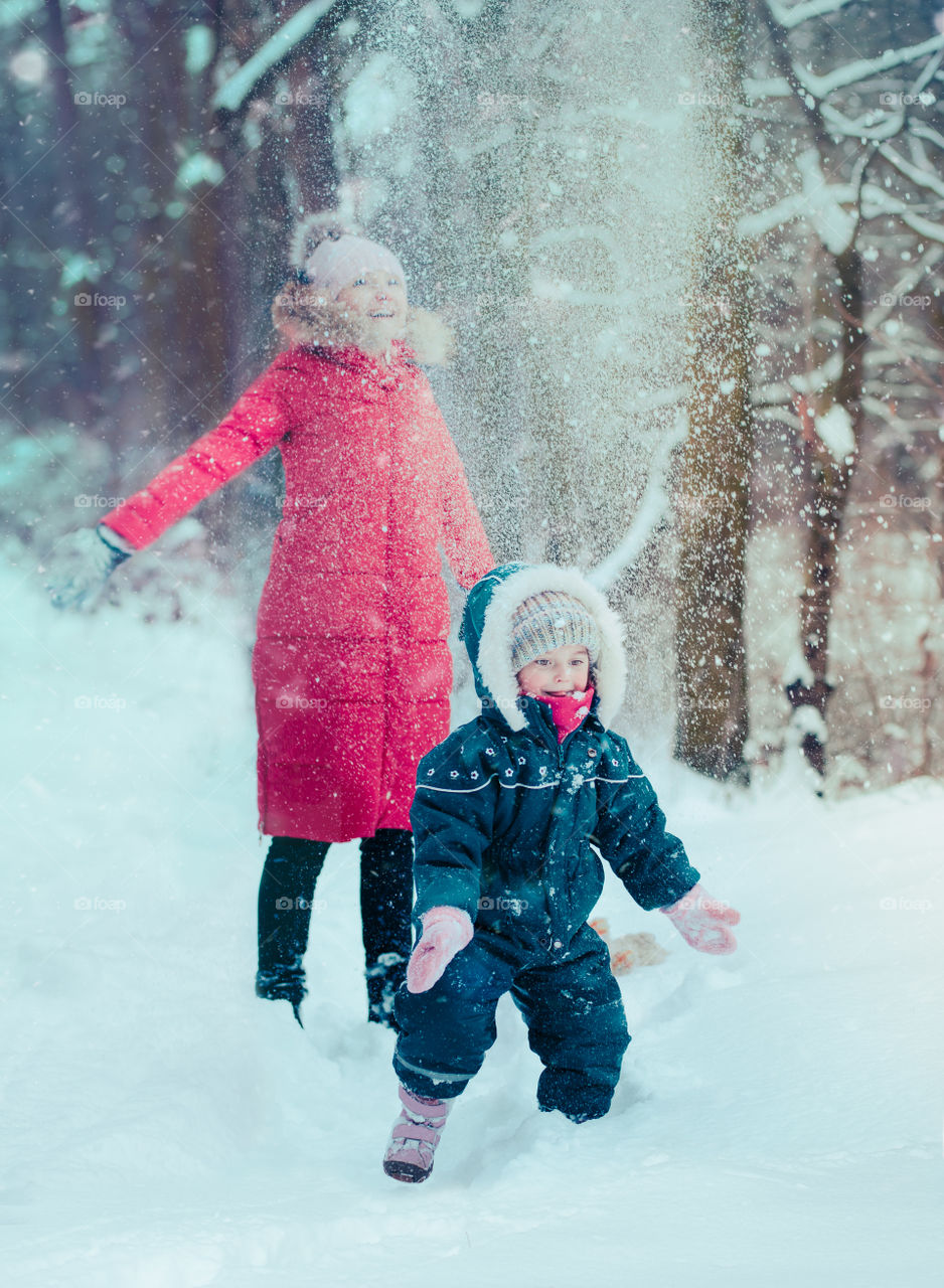 Mother is playing with her little daughter outdoors on wintery day. Woman is throwing snow on her child. Family spending time together enjoying wintertime. Woman is wearing red coat and wool cap, toddler is wearing dark blue snowsuit
