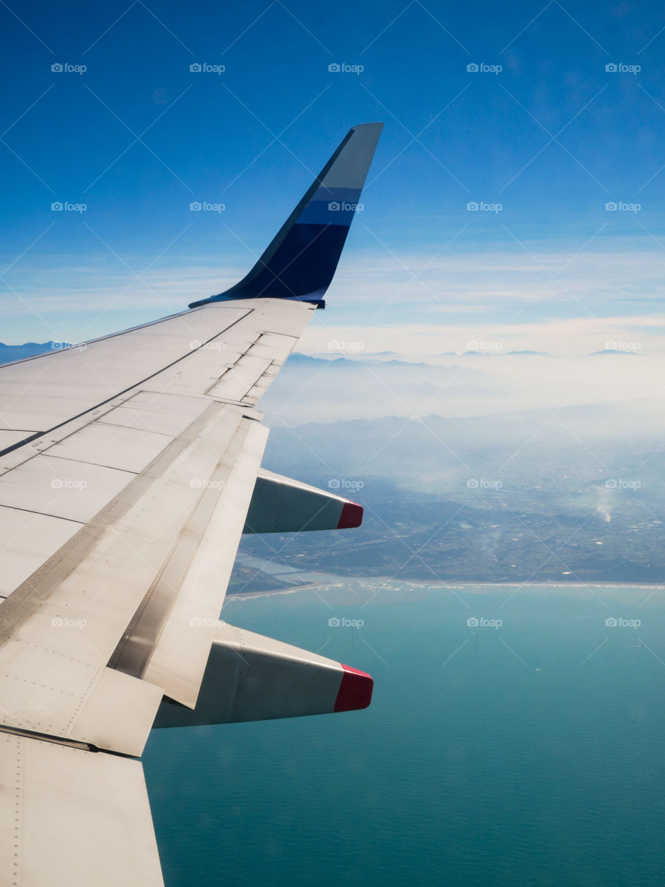 A day shot of a plane wing flying over the ocean a city and mountains. Nice contrasting blue colors all over