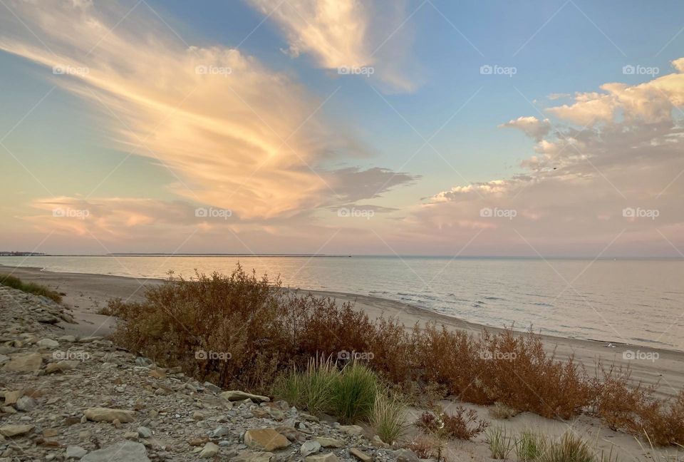 Liquid Fresh water Lake Eerie on Cedar Point shoreline and sandy, rocky beach with purple, pink and blue sunset sky divided by clouds. Beach grass and shrubs in foreground