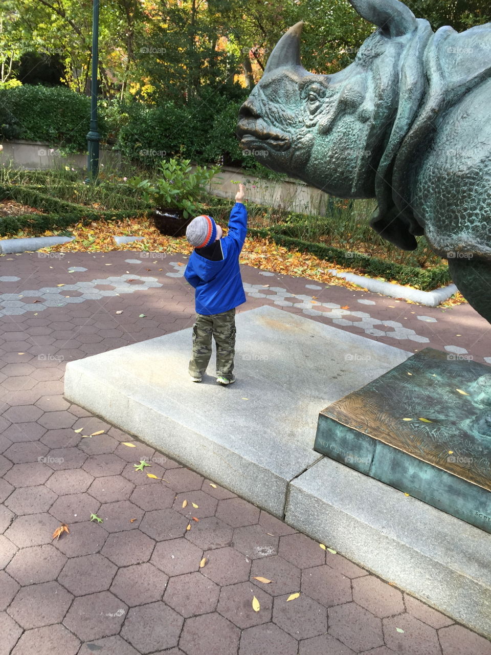Little boy in a blue jacket and striped hat reaching up to the face of the huge rhino rhinoceros statue at the NYC Bronx Zoo in New York City. 