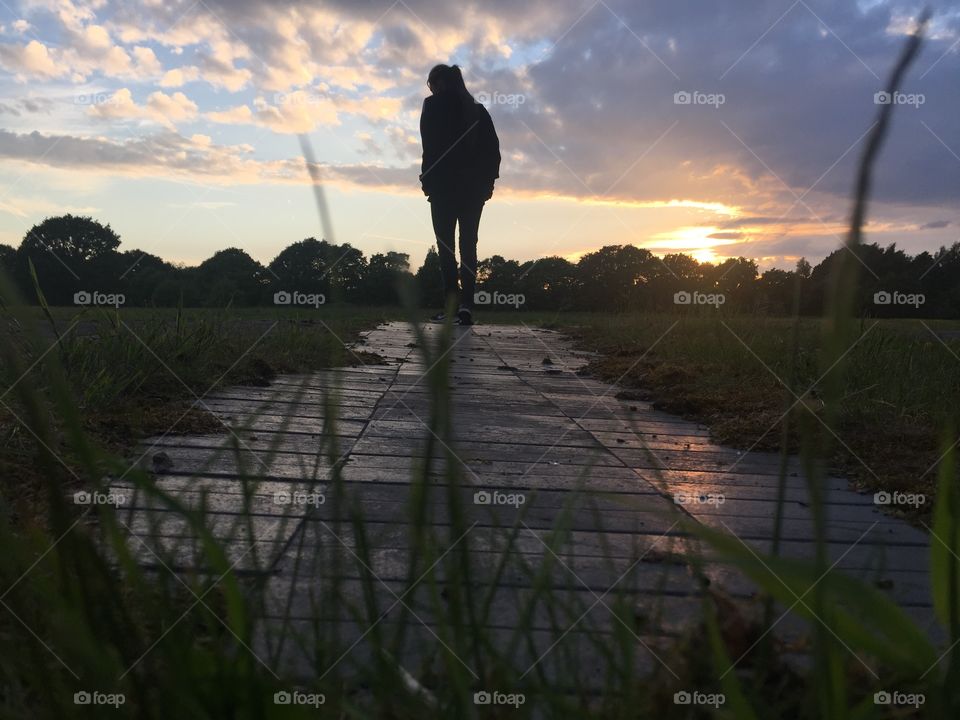 Girl walking away from the camera whilst the grass consumed the bottom of the image. The light reflects off the path whilst a beautiful sunset develops in the background sky
