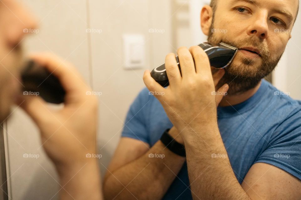 Millennial man using shaving machine to cut off his beard.