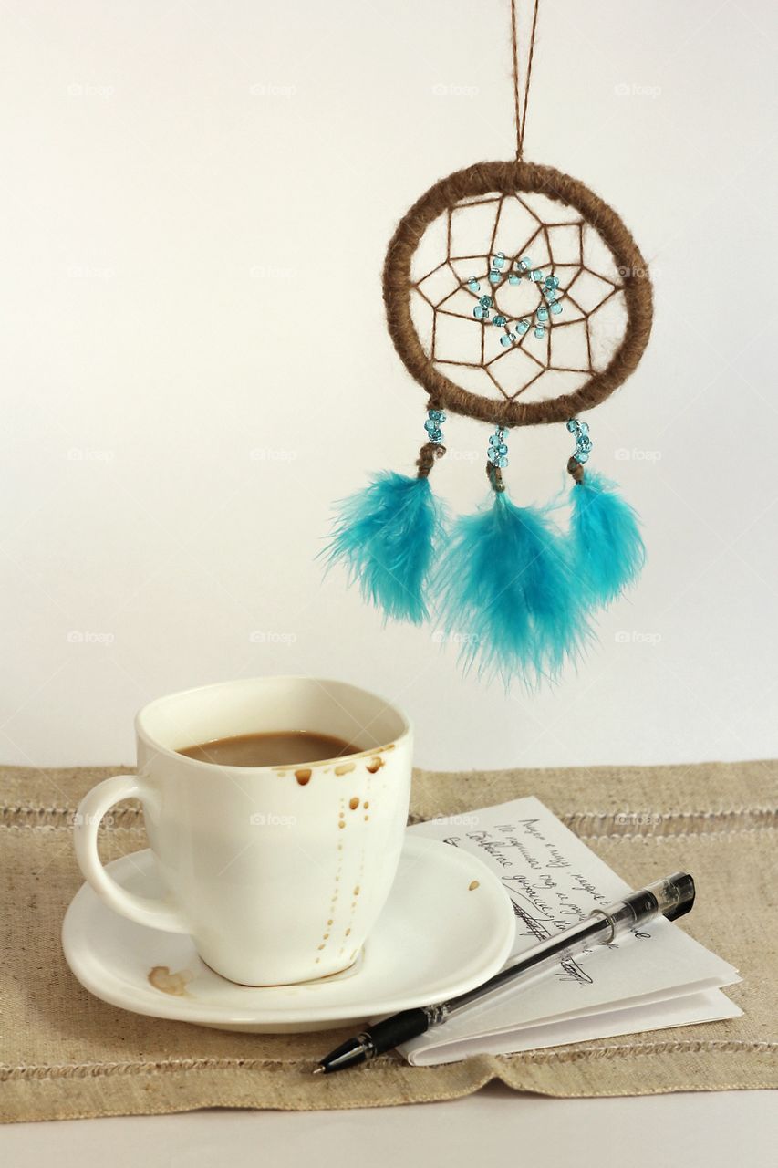 Still life of coffee in a white cup, dream catcher with blue beads and feathers and unfinished verses