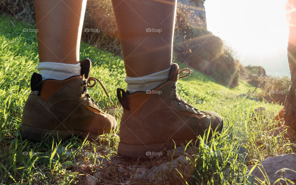 Feet with hiking boots on standing on a rocky grass surface towards the Sun.
