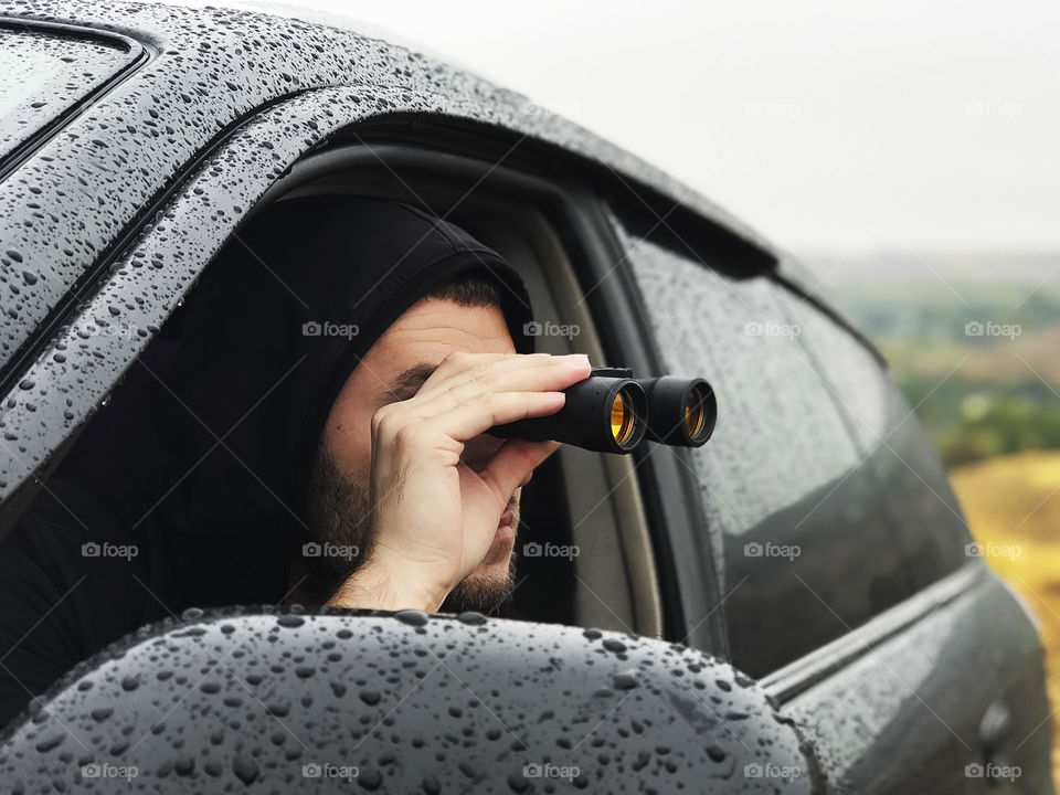 Young man watching through the binoculars out of the car during the rainy day 