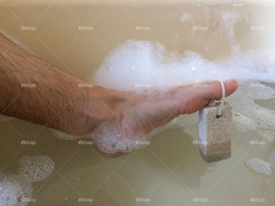 Male foot covered in soap suds in bath with pumice stone hanging from toe