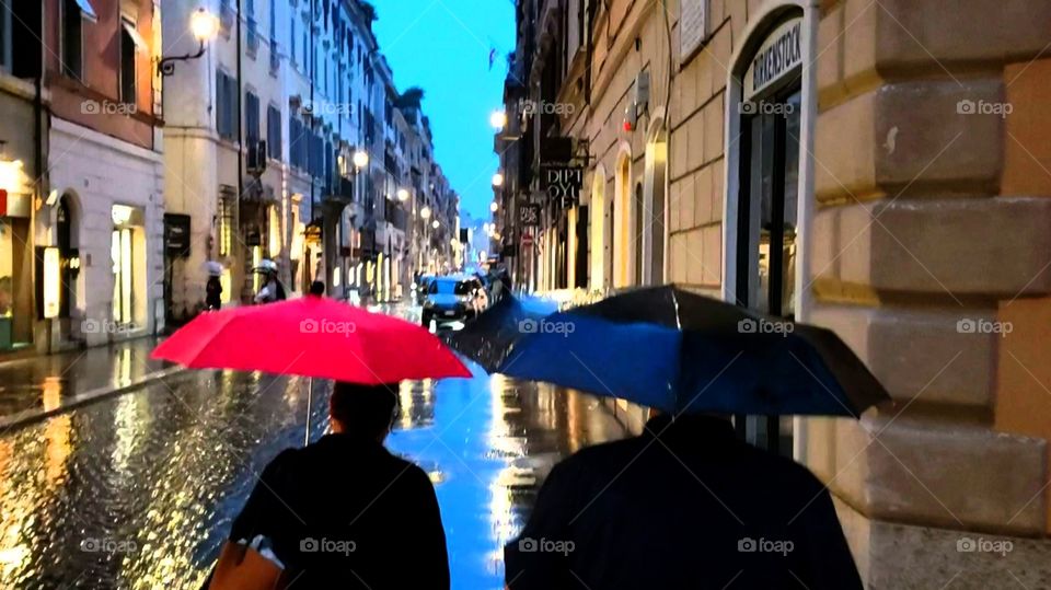 Two people walking in the rain, with red and blue umbrellas, down a street in Rome, Italy, during evening with streetlights reflecting on the side walk. Romantic.