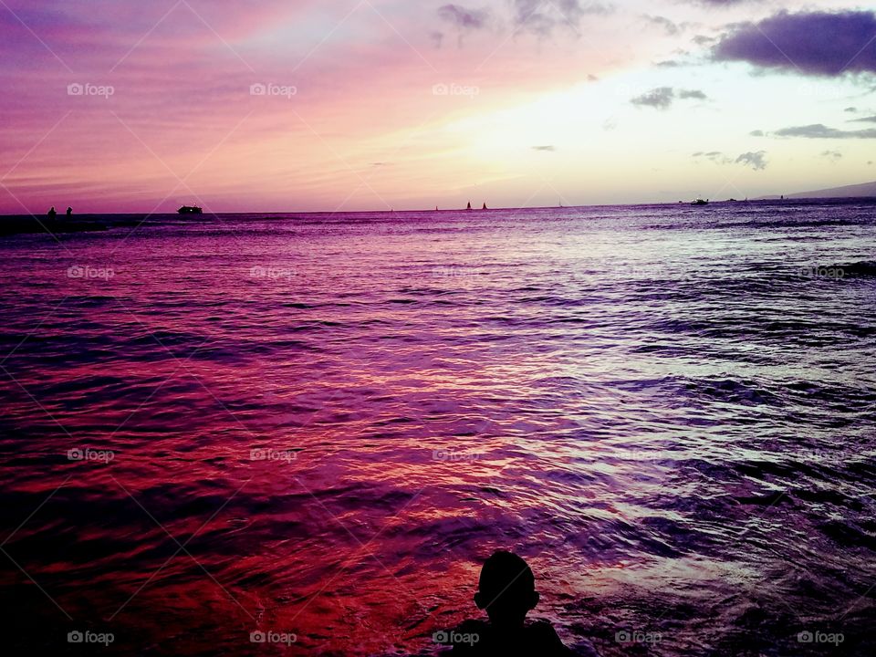 silhouette of young boy in front of beautiful shades of red and purple on the ocean during sunset at Waikiki Beach