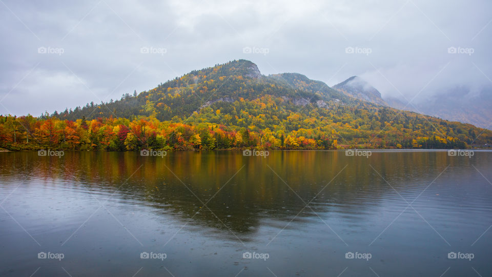 Echo Lake in rain. Clouds and mist lingering with mountains in morning. Reflection in water of mountains and colorful yellow and red autumn leaves. Franconia Notch, NH. New England fall foliage.
