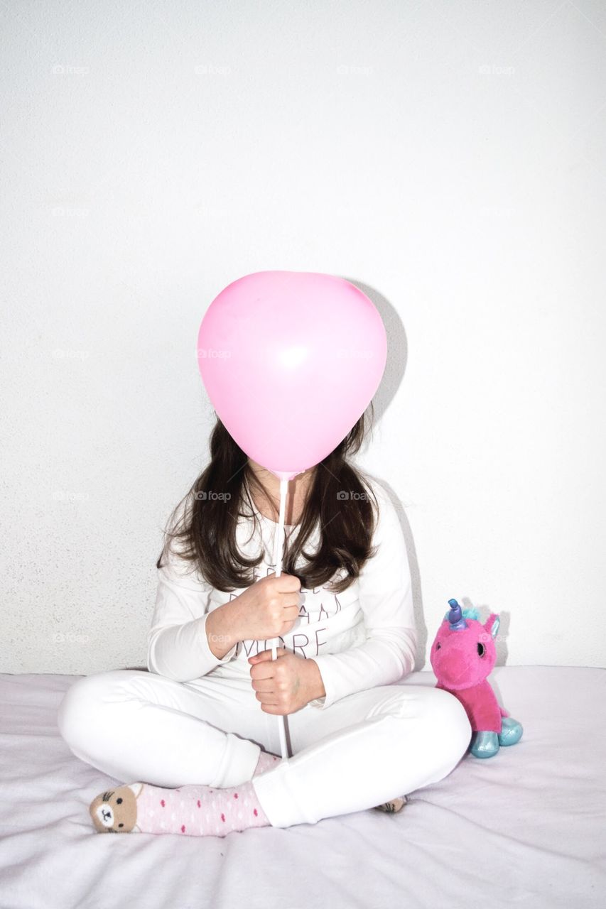 Little girl sitting next to a unicorn blue and pink while holding a pink ballon with white stick.
