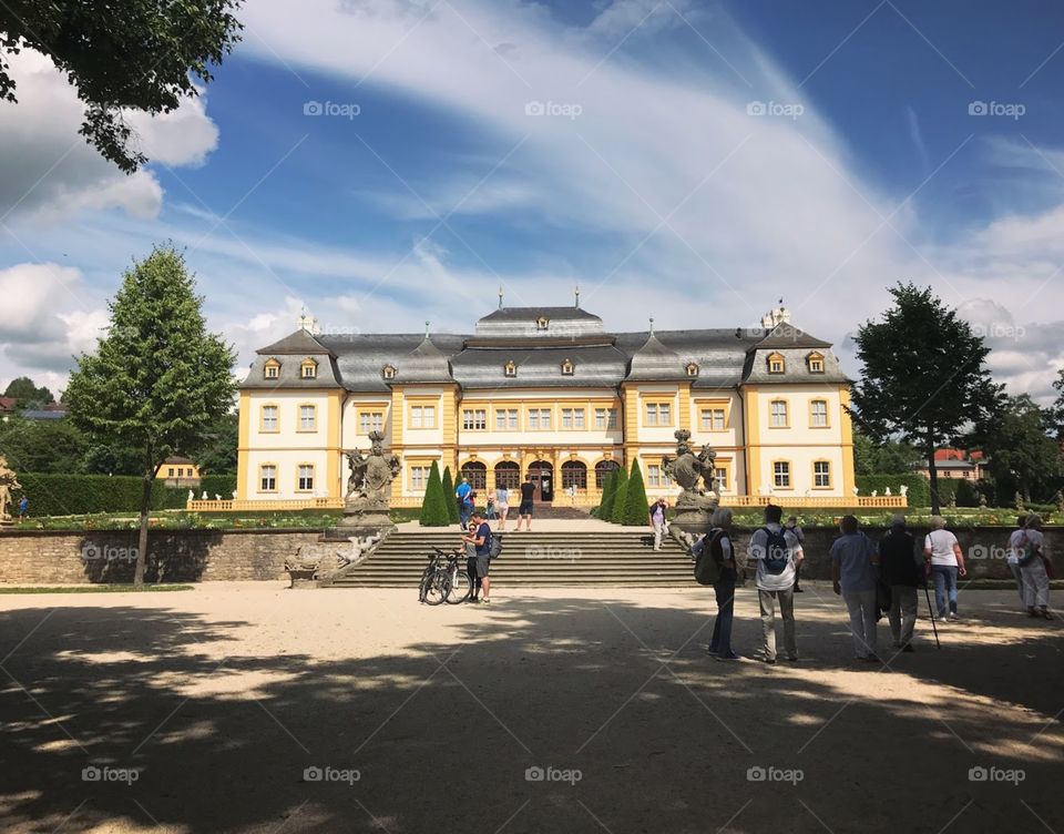 a sleek castle that stands out against a blue sky and white clouds, many people standing to admire the beauty and take pictures, good weather