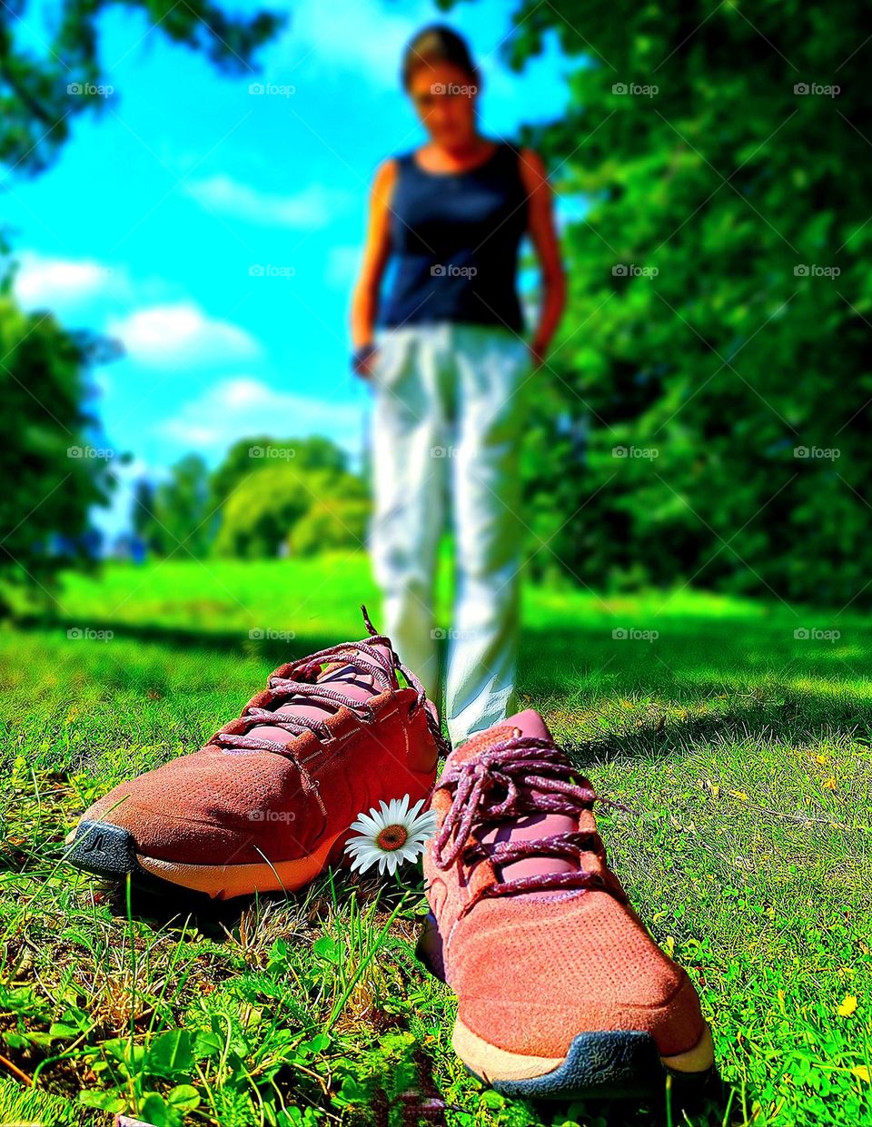 Spring. Burgundy sneakers stand on the green grass near a daisy. In the background is a silhouette of a woman